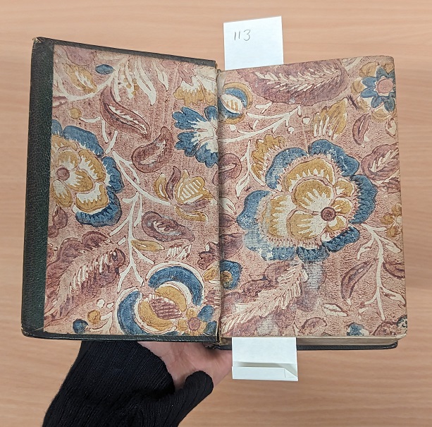 A hard back book is open at the first page revealing the decorated end pages. The pages have been block printed with a floral pattern.