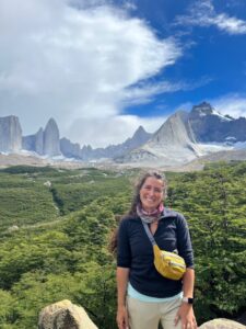 A woman smiles for the camera. She stands in a grand landscape, with green hills and tall sharp mountains further back. 
