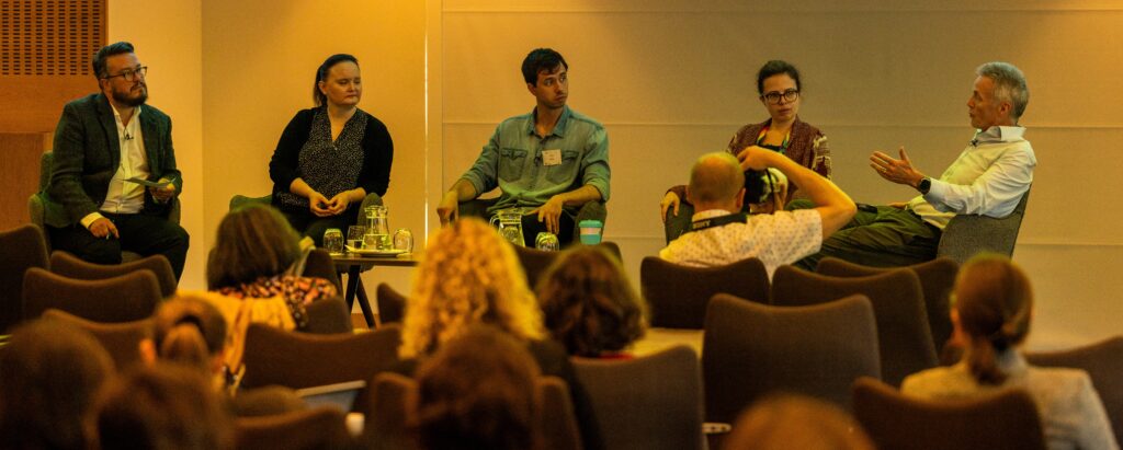 Panel host Dominic and panel speakers Rowena, Nick, Katie, and Malcolm sit on a stage in front of the conference audience