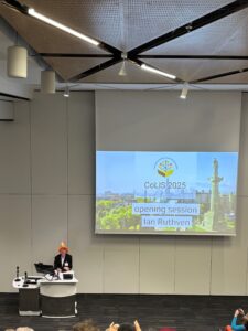 Conference chair, Ian Ruthven, giving a welcome speech to the conference attendees. He is wearing a hat that looks like a traffic cone, in reference to a statue in Glasgow that has a traffic cone on it. An introdcutory slide from a presentation is on the screen.